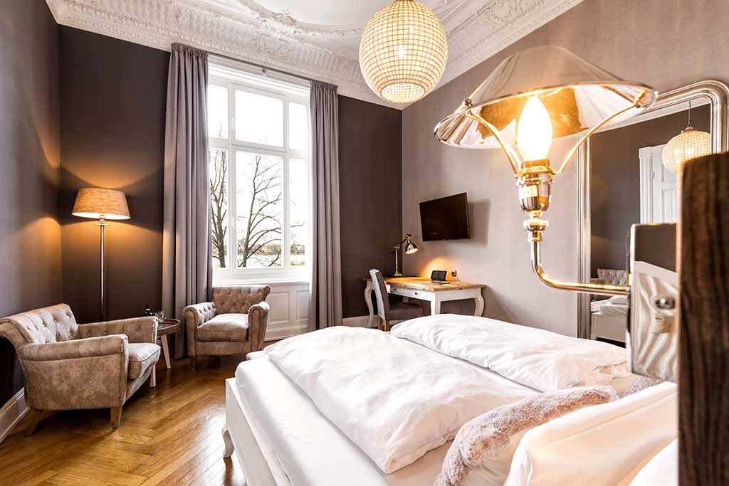 Helles, elegantes Schlafzimmer mit hohen Fenstern, modernem Kronleuchter, grauen Wänden, einem Doppelbett mit weißer Bettwäsche, zwei beigen Sesseln, einem Schreibtisch mit Stuhl, einer Lampe und einem Fernseher, mit klassischem Stuck an der Decke.