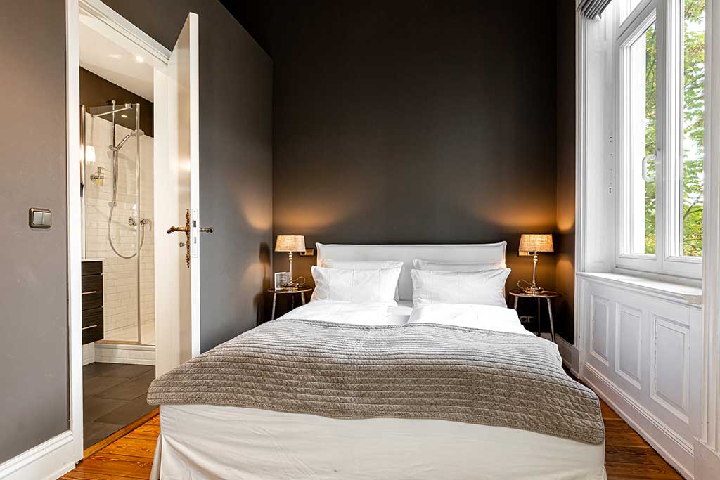 Ein modernes Schlafzimmer mit dunklen Wänden, einem Doppelbett mit weißer Bettwäsche und grauer Decke, Nachttischen mit Lampen, Holzboden, einem Fenster mit Blick nach draußen und einer offenen Tür zu einem hellen Badezimmer.