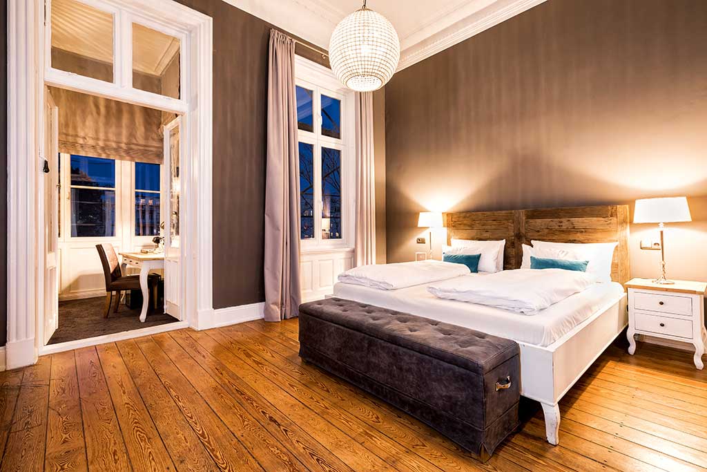 Ein stilvolles Schlafzimmer mit Holzfußboden, einem Doppelbett mit weißer Bettwäsche und blauen Kissen, zwei Nachttischen mit Lampen, einer grau gepolsterten Bank und einem durch hohe Fenster und Vorhänge einsehbaren Nebenraum.